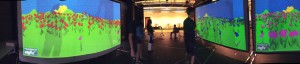180° panoramic image of an installation of Sow/Reap. The image depicts five people standing before three large screens. The screens have pink, blue, and red flowers in a landscape. The flowers are "planted" by the gestures of the people.